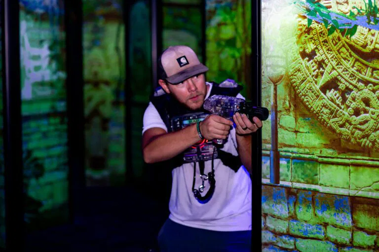 A man grips a gun while standing before a wall at The Rush Funplex, evoking a sense of suspense or intensity.