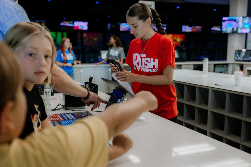 A woman hands a toy to a child at The Rush Funplex bowling alley, surrounded by colorful bowling lanes and decorations.